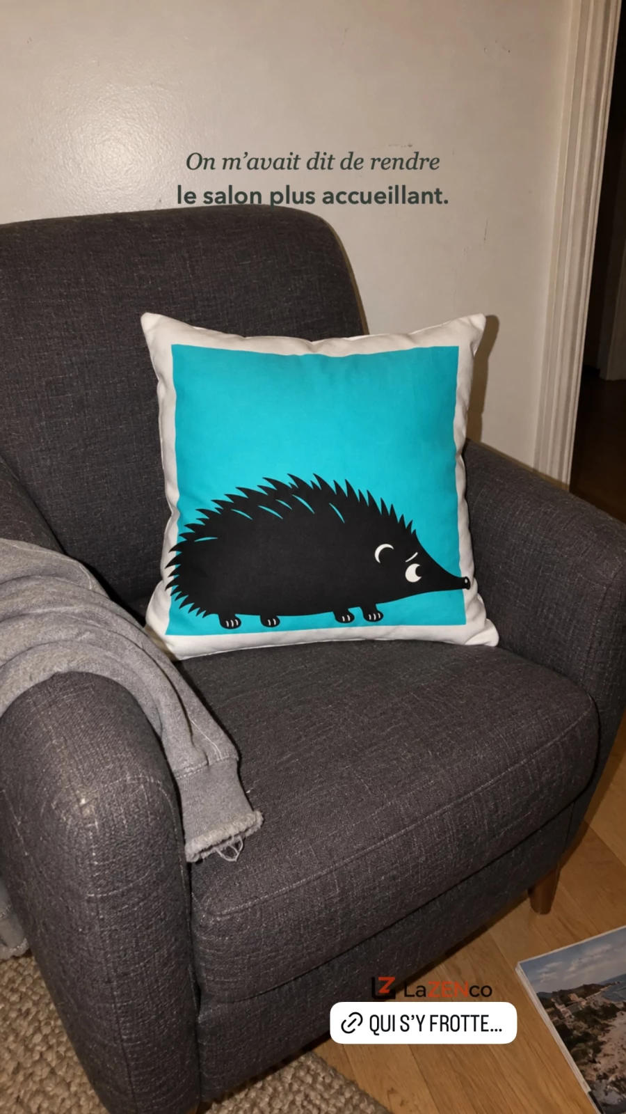Coussin blanc au carré turquoise et au hérisson noir piquant, sur un fauteuil gris.